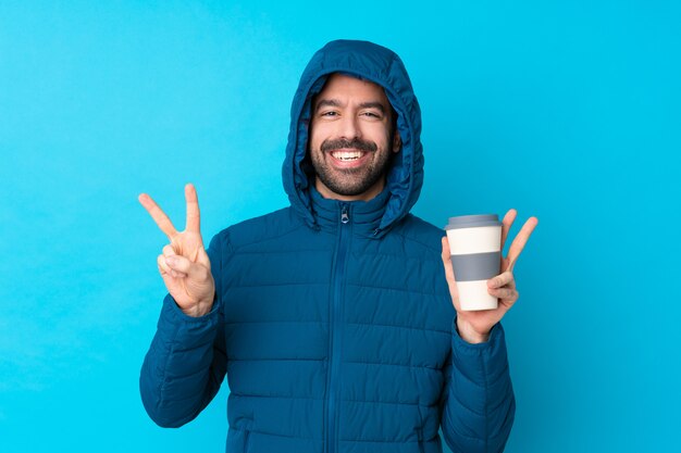 Homem vestindo jaqueta de inverno e segurando um café para viagem sobre parede azul isolada, mostrando sinal de vitória com as duas mãos