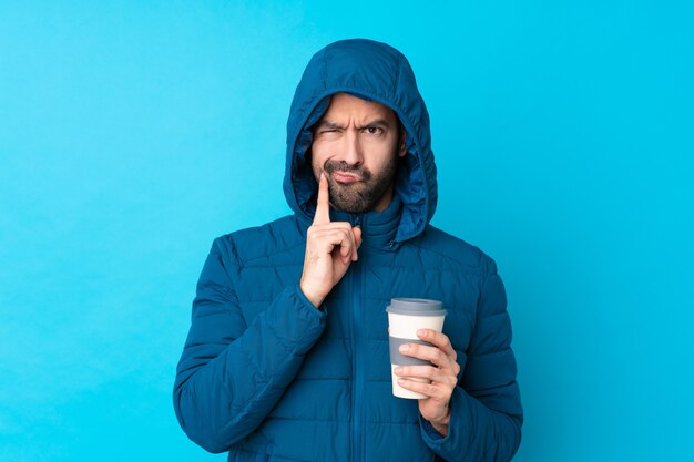 Homem vestindo jaqueta de inverno e segurando um café para viagem sobre parede azul isolada, olhando de frente