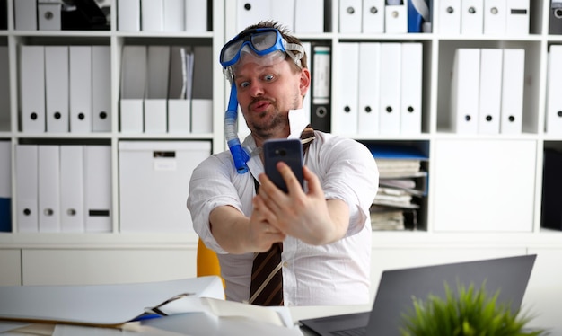 Homem vestindo terno e gravata em óculos e snorkel faz selfie com celular no escritório pronto para tirar o retrato Contar dias para sair do dia anual de folga workaholic liberdade diversão turismo resort ideia conceito