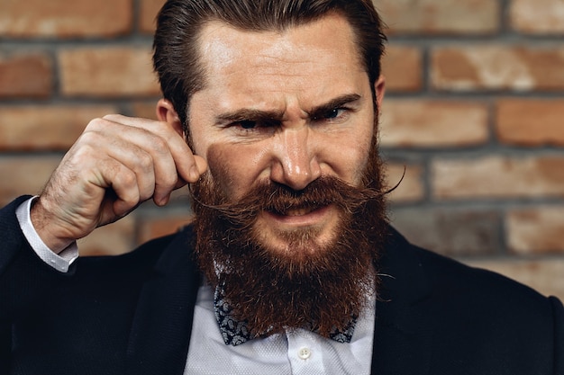 homem vestindo um terno, bigode de hussardo e barba e posando contra uma parede de tijolos.