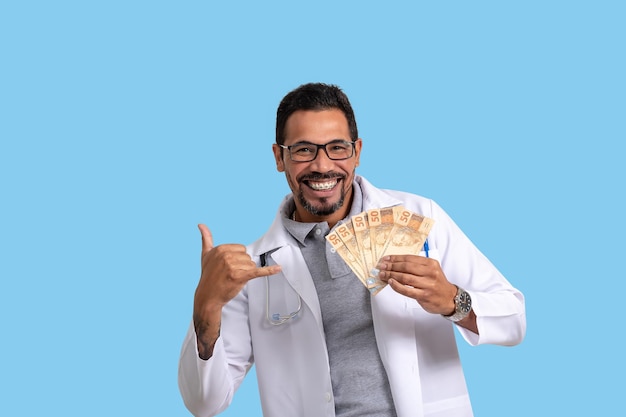 Foto homem vestindo uniforme médico segurando dinheiro brasileiro, sorrindo, expressão autoconfiante, em azul bac