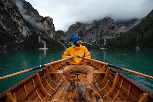 Homem viajante com chapéu azul e capa de chuva amarela, navegando em um barco de madeira pelo belo lago Di Braies nas montanhas Dolomitas.