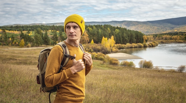 Homem viajante com um chapéu amarelo e um suéter com uma mochila está em frente ao rio, a floresta dourada de outono e as colinas. Conceito de liberdade, viagens, caminhadas