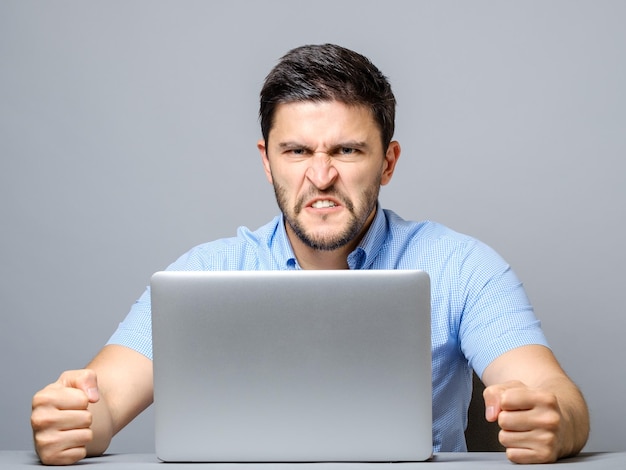 Homem zangado sentado à mesa com laptop sobre fundo cinzento