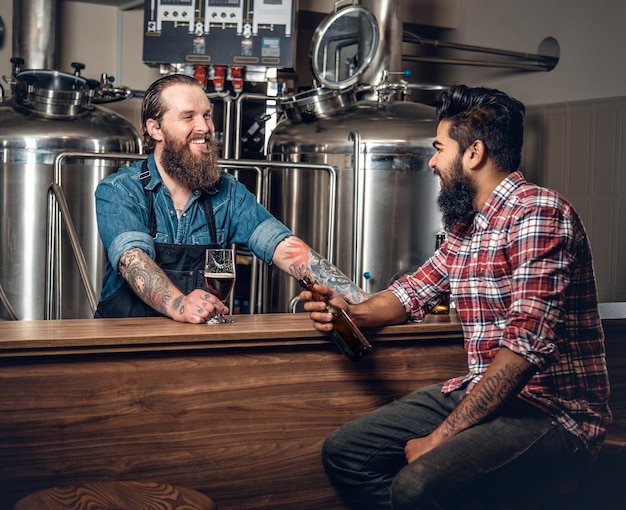 Foto homens caucasianos e indianos barbudos tatuados apresentando cerveja artesanal na microcervejaria.