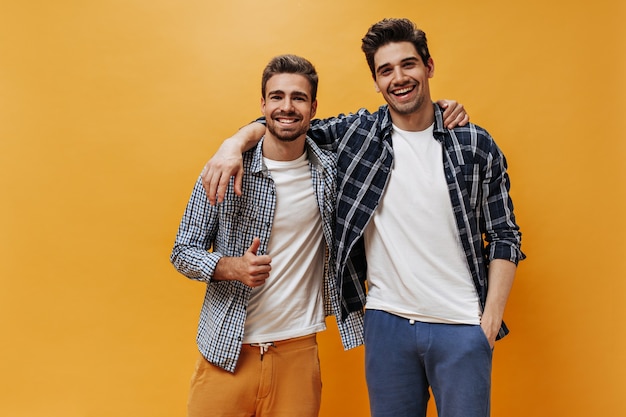 Foto homens charmosos em camisas xadrez e camiseta branca sorriem na parede laranja. cara barbudo legal mostra o símbolo da paz.