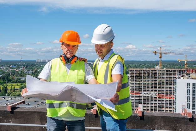 Homens com capacete branco seguram um plano de construção e inspecionam o canteiro de obras