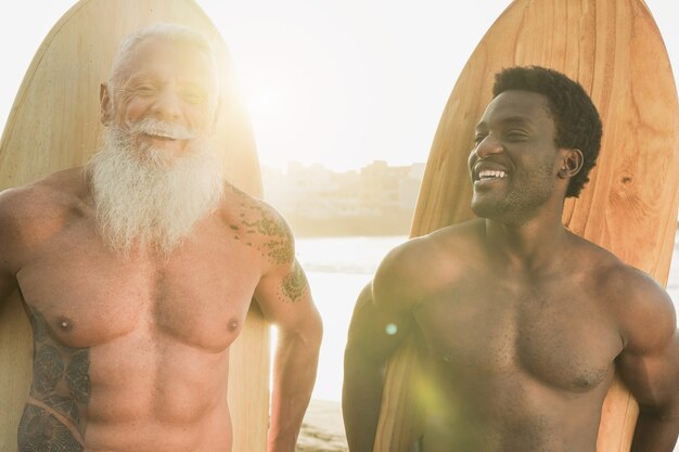 Homens de várias gerações na praia com prancha de surf - Surfistas multirraciais ao pôr do sol ao ar livre