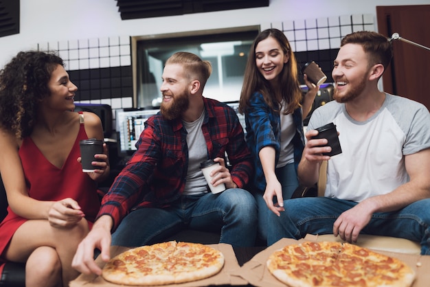 Homens e duas mulheres no estúdio de gravação estão comendo pizza.