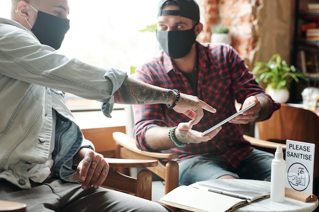 Homens jovens com máscaras de pano sentados no saguão e discutindo um novo aplicativo no tablet