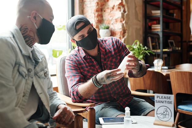 Homens jovens com máscaras pretas sentados em um café aconchegante e verificando o desinfetante enquanto lêem o rótulo.