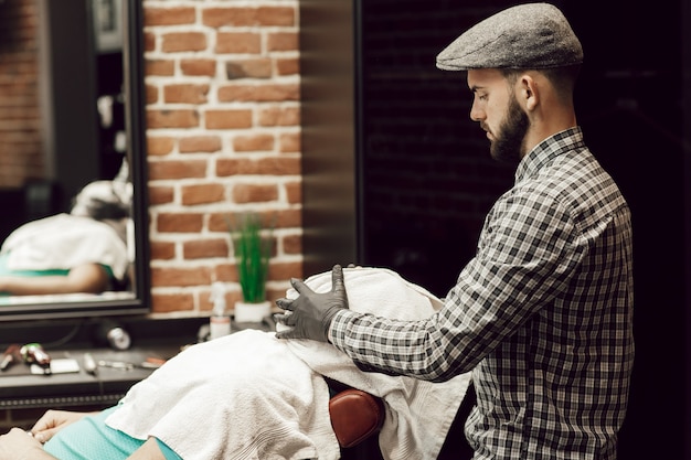 Homens jovens em uma barbearia. O cabeleireiro enxuga o rosto da cliente com uma toalha após raspar a barba e cortar o cabelo.