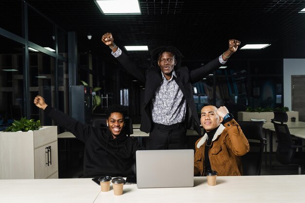 Homens multiétnicos felizes e alegres levantam as mãos e os punhos e se alegram em ganhar no cassino online no laptop Formação de equipes de africanos negros e programadores asiáticos