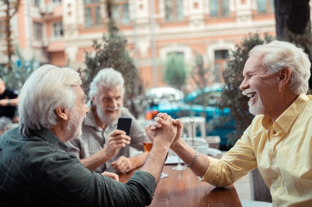 Homens rindo. homens alegres de cabelos grisalhos rindo enquanto lutam contra a queda de braço sentados do lado de fora do bar