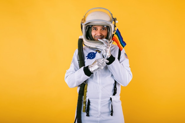 Homosexueller männlicher Kosmonaut im Raumanzug und im Helm, der lgtbi Regenbogenfahne auf gelber Wand hält.