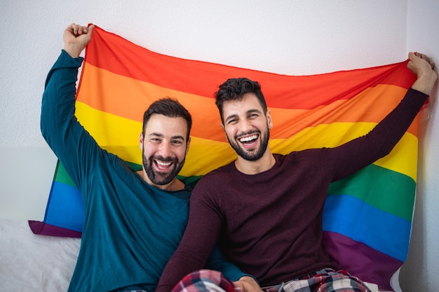 Homosexuelles Paar, das lgbt Regenbogenfahne drinnen auf Bett zu Hause hält - Fokus auf rechtes Manngesicht