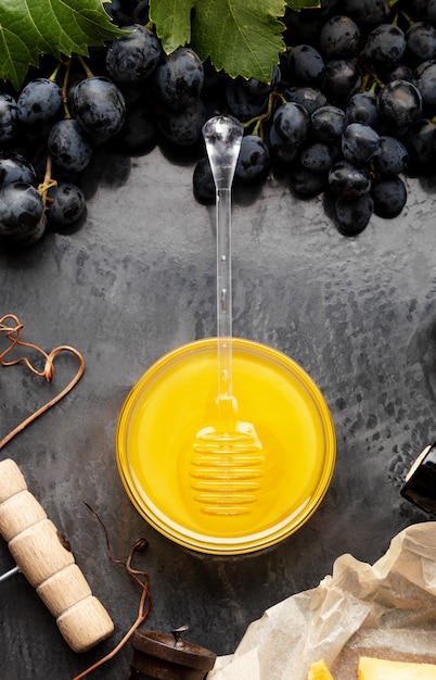 Honig im Glas mit Honiglöffel auf dunklem Steinbrett mit Weinkäse-Korkenzieher im schwarzen saftigen Traubenrahmen. Köstlicher gelber Bienenhonig im Snackteller auf schwarzem Tisch. Ansicht von oben. Vertikales Foto.