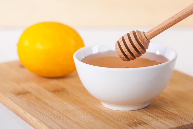 Honig in einer weißen Keramikschale mit Honigschöpflöffel und Zitrone auf einem hölzernen Küchenbrett.