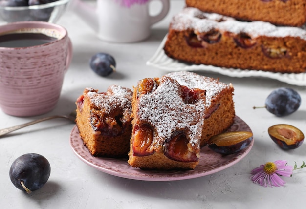 Honigkuchen mit Pflaumen, bestreut mit Puderzucker und Tasse Kaffee auf hellgrauem Tisch