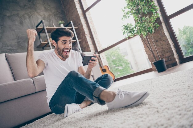 Hooray! Foto de mestiço louco sentado no chão, encostado no sofá, segurando o telefone, lendo notícias positivas inesperadas assinantes do blog vestem roupa casual na sala de estar dentro de casa