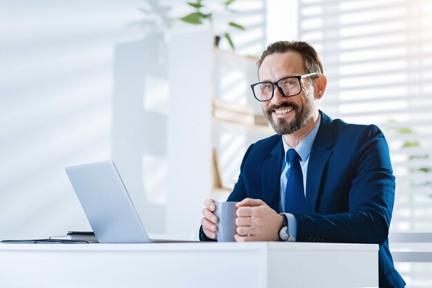 Hora de cafe. Alegre empresario alegre positivo sentado en la mesa mientras sostiene la taza y sonriendo