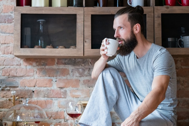 Hora de cafe. Contemplación matutina. Intención. Joven hipster barbudo con taza blanca sentada en la encimera de la cocina.