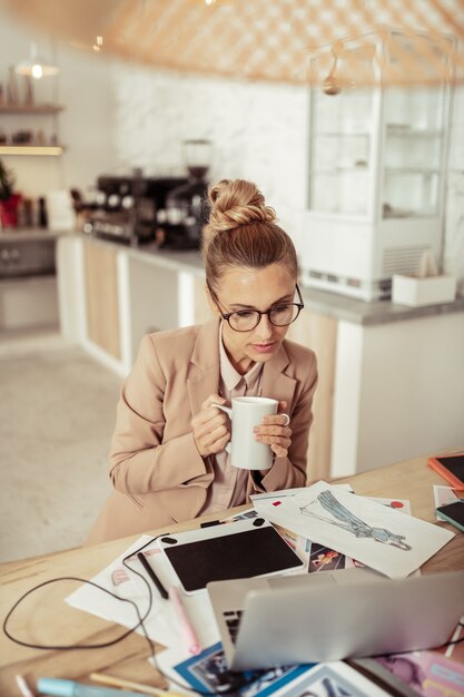 Hora de cafe. Hermosa diseñadora sentada en la mesa tomando café y mirando sus bocetos.