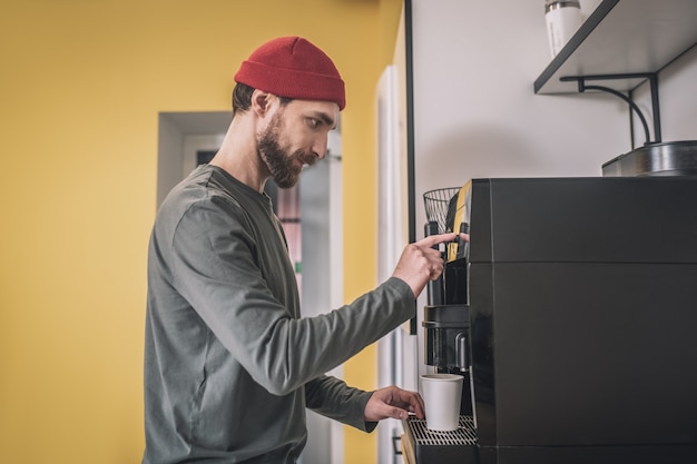 Hora de cafe. Hombre con un sombrero rojo de pie cerca de la máquina de café