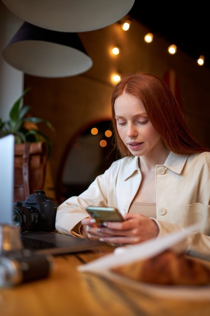 Hora del café, mujer con teléfono móvil y portátil en la cafetería, trabajando. Retrato de vista lateral de mujer pelirroja linda en ropa casual sentado detrás de la mesa, croissant en la mesa