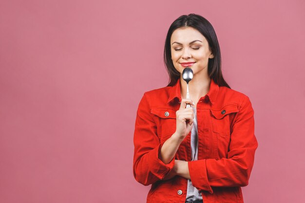 ¡Hora de cenar! Retrato de la mujer hermosa joven que lleva la camisa casual roja, sosteniendo la cuchara aislada sobre fondo rosado.