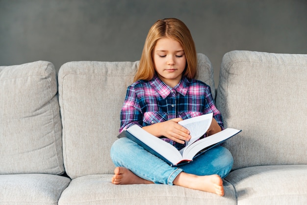 Hora de los cuentos de hadas. Hermosa niña leyendo un libro mientras está sentado en el sofá en posición de loto en casa