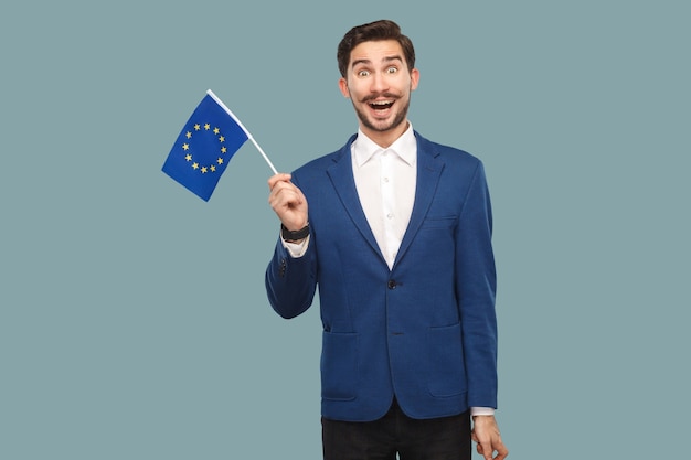 É hora da Europa. Bonito empresário surpreso de jaqueta azul e camisa branca, segurando a bandeira da União Europeia e olhando para a câmera com rosto maravilhado. Interior, estúdio tiro isolado em fundo azul claro.