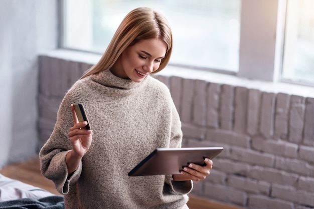 Hora das compras. Jovem mulher muito alegre usando cartão de crédito e tablet enquanto relaxa e está sentado no quarto.