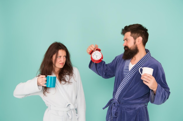 Hora de acordar e ter um bom dia Marido de cara segura café e relógio Todas as manhãs começam com café Casal em roupões de banho com canecas Homem com barba e mulher sonolenta desfrutam de café ou chá da manhã