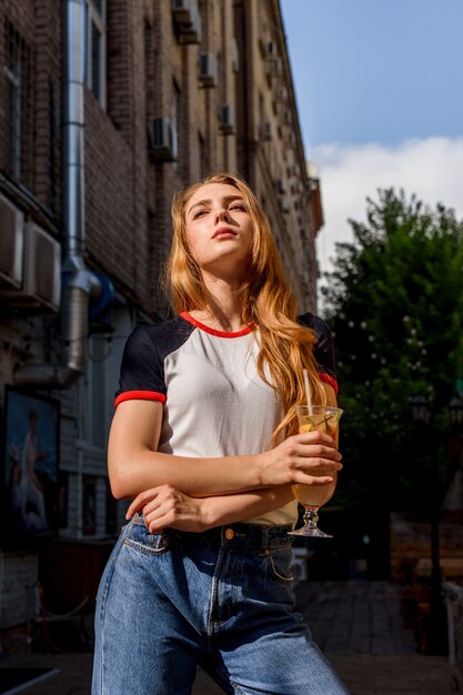 É hora de beber um coquetel. Uma mulher muito bonita bebe um coquetel. Loira natural em camiseta e jeans