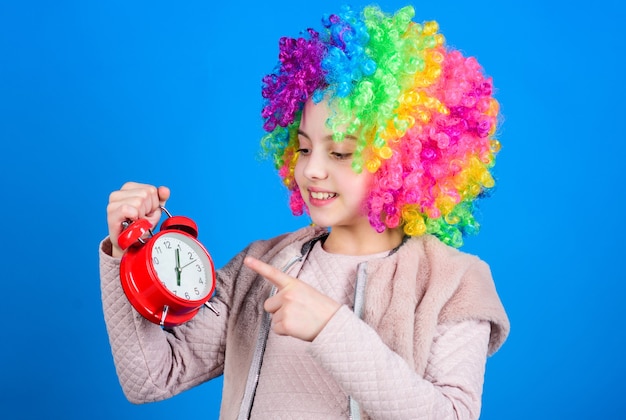 É hora de descanso. Menina feliz com cabelo de peruca colorida apontando para o despertador para a hora exata. Conceito de tempo. Praticar uma boa gestão do tempo.
