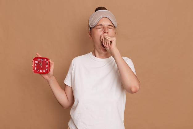 Hora de dormir Foto interior de bocejando jovem adulta segurando despertador isolado em fundo marrom mantém os olhos fechados cobrindo a boca com o punho acorda de manhã cedo sem dormir