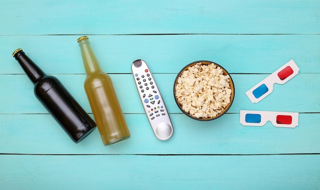 Hora de filme. Garrafa de cerveja, controle remoto da tv, óculos 3d e tigela de pipoca sobre fundo azul de madeira. Vista do topo