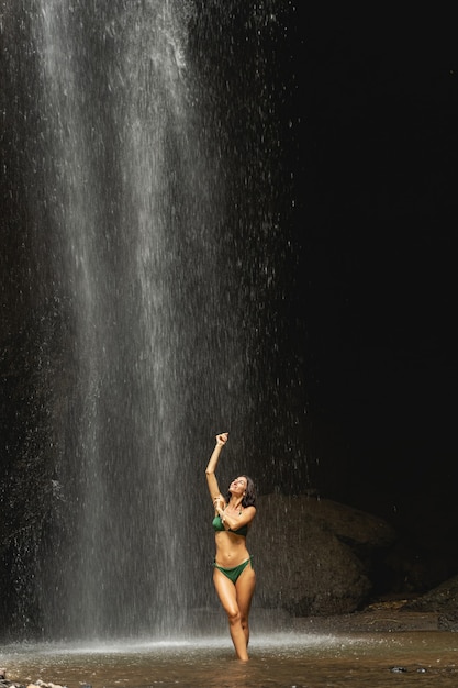 É hora de nadar. Mulher morena encantada positiva em pé sob a cachoeira lavando o corpo, lugar exótico