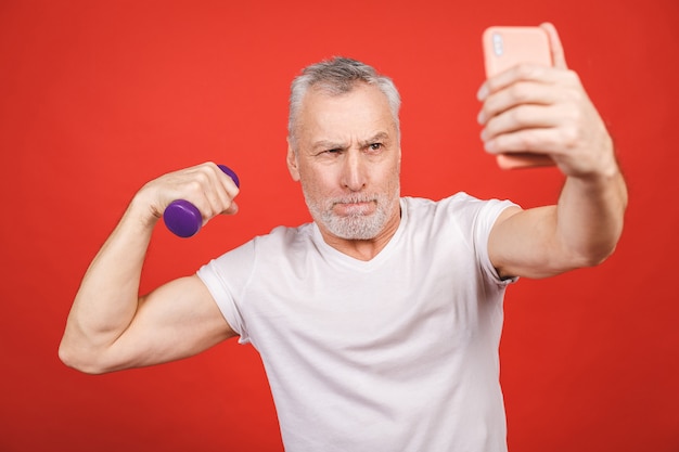 É hora de tirar uma selfie! Retrato do close-up de um homem sênior que exercita com halteres. Usando o telefone