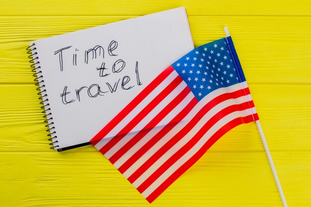É hora de viajar para os EUA. Bandeira dos EUA e o bloco de notas na mesa de madeira amarela no fundo.