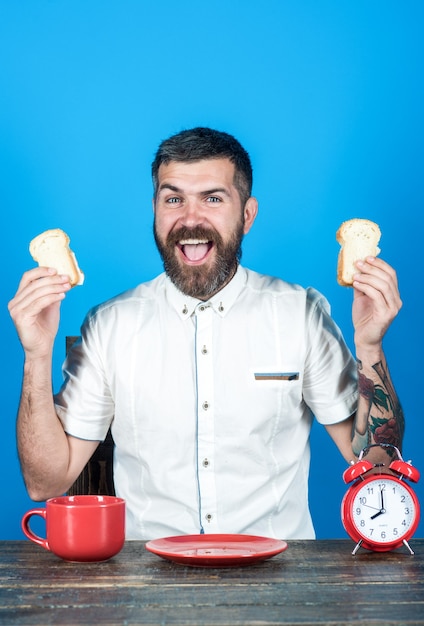 Foto hora del desayuno. desayuno por la mañana. hombre barbudo comiendo deliciosas tostadas. hombre guapo satisfecho con camisa blanca sentado a la mesa y comiendo. el hombre barbudo come de la placa roja. hora de cafe.