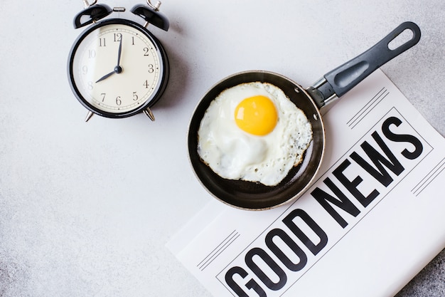 Hora del desayuno. Huevos fritos en una sartén con un reloj despertador y un periódico sobre una mesa de color gris claro con un fondo de textura.