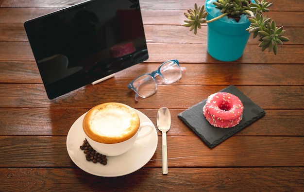 Hora do café da manhã com cappuccino quente e glacê rosa donut tablet digital para as últimas notícias bom dia na mesa de madeira rústica