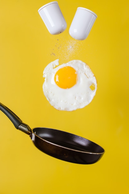 Foto hora do café da manhã. um ovo frito e uma frigideira com saleiros levitam no ar em um fundo amarelo