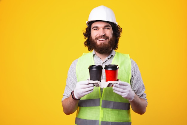 Hora do café para a melhor equipe de trabalho. engenheiro barbudo alegre usando capacete e segurando duas xícaras de café para viagem