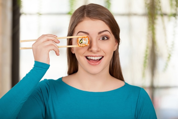 Hora do sushi! Mulher jovem e bonita segurando sushi na frente do olho e sorrindo enquanto está sentada no restaurante