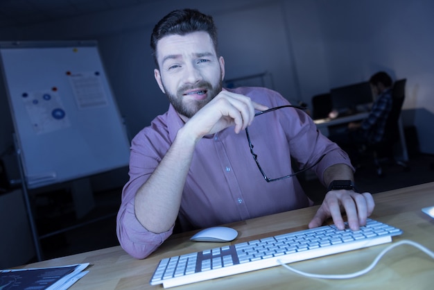 Hora extra. Homem de TI cansado e bonito tirando os óculos e descansando do trabalho enquanto está sentado em frente ao computador