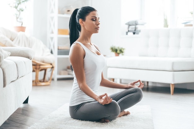 Hora de hacer yoga. Atractiva mujer joven haciendo ejercicio y sentado en posición de loto de yoga mientras descansa en casa