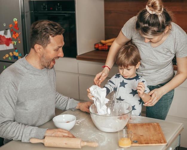 Horas felices de familia joven, papá se ríe. Mamá e hijo se dedican a hornear, sentados a la mesa en la cocina. Lección de horneado en casa, trabajo en equipo, gente feliz, niño pequeño involucrado en el proceso